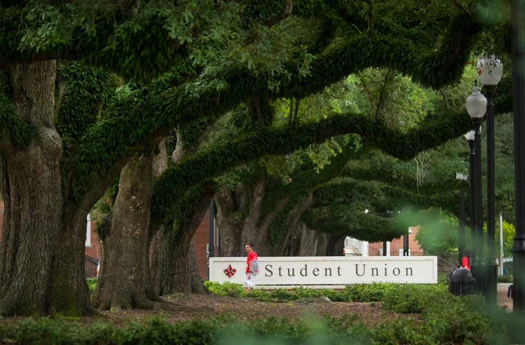 Student walking in front of the University of Louisiana at 69传媒 Student Union sign under a canopy of oak tree branches